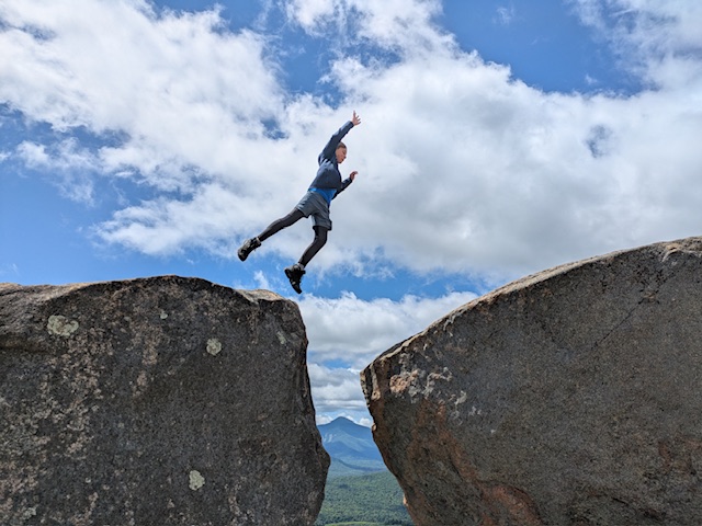 Image of a young boy leaping between 2 boulders on a summer day. Sky is blue with fluffy clouds. Between, the boulders, in the distance you can see a far of mountain. 