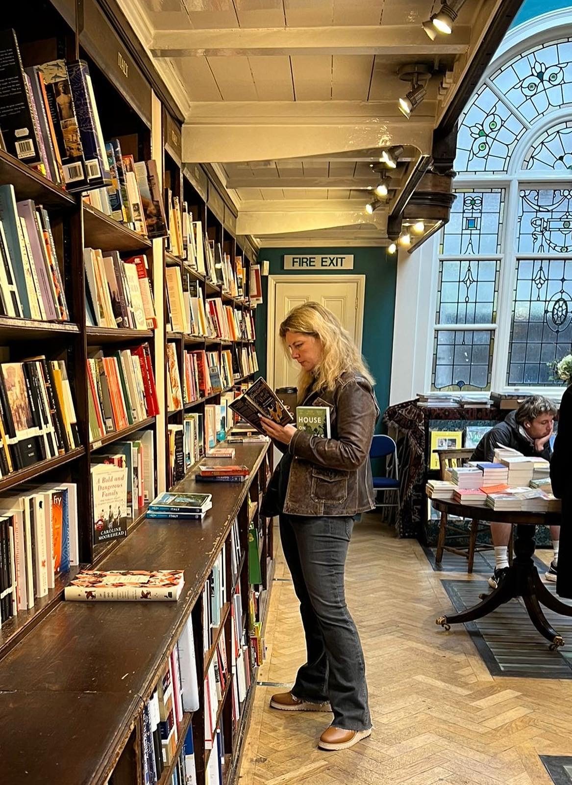 A woman stands in a library for bookstore facing a long shelf filled with books. In her arms she holds a stack while simultaneously reading one.
